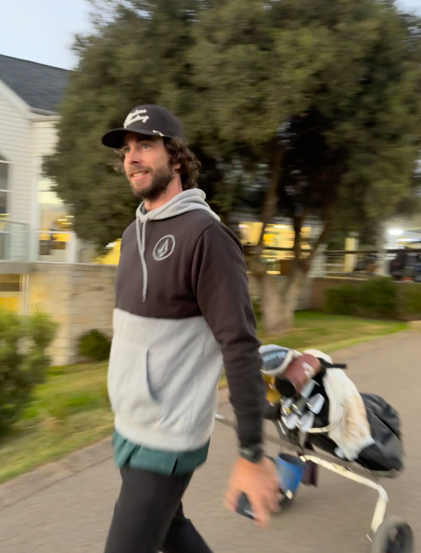 A man walking outdoors while wearing a colorblock hoodie with a dark brown upper section, light gray middle section, and teal hem, along with a black cap and a golf bag on a push cart.